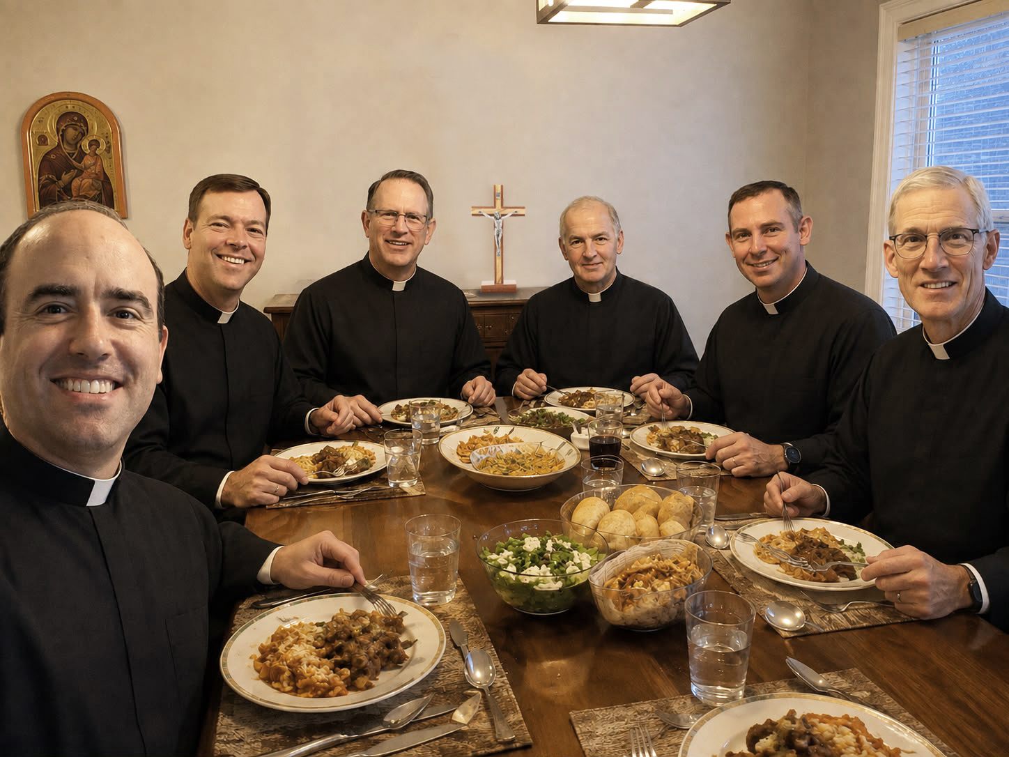 Six priests seated around a wooden dining table sharing a meal, beneath a crucifix and an icon of the Virgin and Child